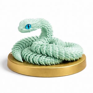 Op de afbeelding: Een mintgroen slangenbeeldje met gedetailleerde schubben en een opvallend blauw oog. De slang is opgerold op een ronde, goudkleurige basis, wat een decoratief stuk creëert. Een uniek woondecoratie-item.
