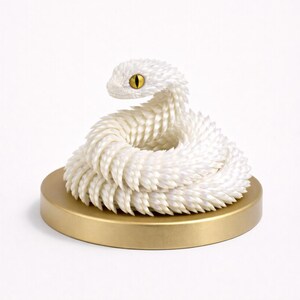 May include: A coiled white snake sculpture with textured scales and a yellow eye, resting on a gold-colored circular base. The snake's head is raised, and its body is arranged in a circular pattern. The sculpture is a decorative item.