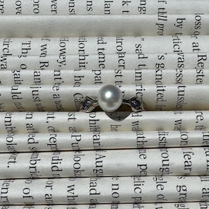 May include: A silver-toned ring featuring a central round white pearl flanked by two small, dark, triangular-cut gemstones set into the metal band. The ring is positioned horizontally against a background of horizontal strips of printed text.