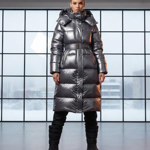 Op de afbeelding: Een zilveren metallic pufferjas met een ceintuur in de taille en een capuchon. De lange jas wordt gedragen met een zwarte broek en zwarte plateau laarzen. De jas heeft een glanzende, reflecterende afwerking en een gewatteerd ontwerp.