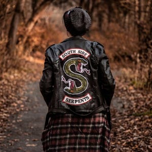 May include: Black leather jacket with a large patch on the back featuring a green snake and the words "SOUTH SIDE SERPENTS". The jacket is worn over a red and black plaid shirt. The person is walking on a path.
