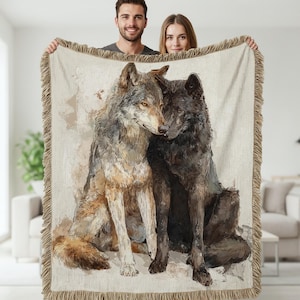 May include: A woven throw blanket featuring a detailed illustration of two wolves, one gray and one black, with a neutral background. The blanket has a fringed edge and is held up by two people.