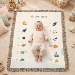 Op de afbeelding: Een baby ligt op een crèmekleurige mijlpaaldeken met de tekst "My first year" en de nummers 1-12 met ruimte-thema illustraties. De deken heeft een gefranjerde rand. De baby draagt een crèmekleurige onesie.