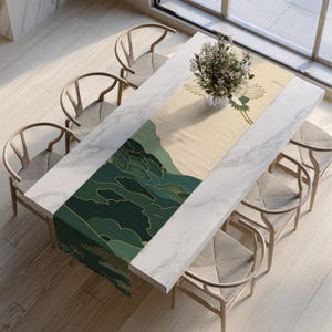 May include: A rectangular table runner with an Asian-inspired design. The runner features a cream-colored background with a green and gold mountain and crane illustration. The runner is placed on a white marble table surrounded by wooden chairs.