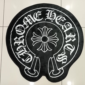 Pode incluir: Tapete redondo preto e branco com o texto "CHROME HEARTS" em fonte g&oacute;tica em torno de um emblema central. O emblema &eacute; uma cruz estilizada com um centro circular. O tapete tem uma textura macia e felpuda.