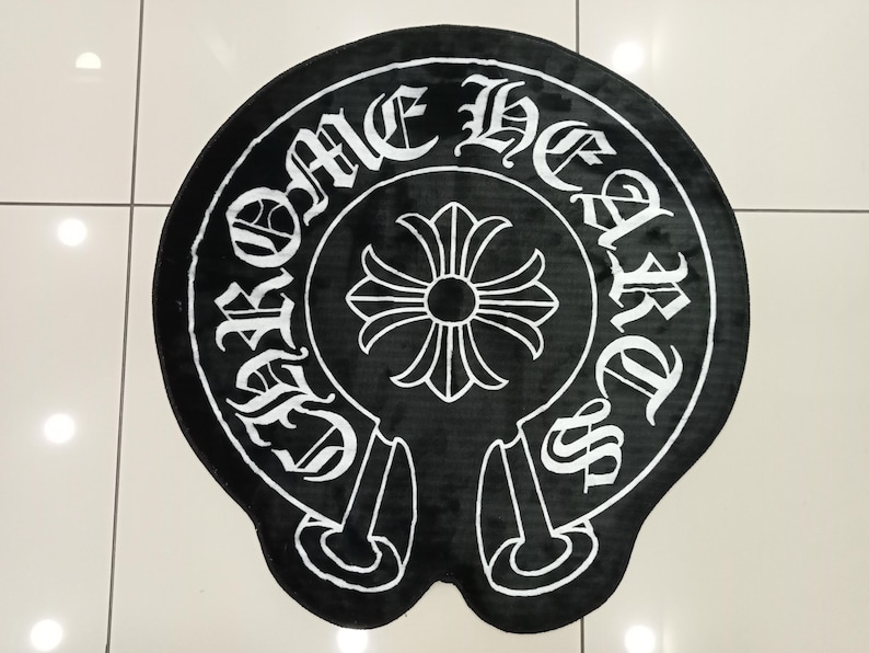 Pode incluir: Tapete redondo preto e branco com o texto "CHROME HEARTS" em fonte g&oacute;tica em torno de um emblema central. O emblema &eacute; uma cruz estilizada com um centro circular. O tapete tem uma textura macia e felpuda.