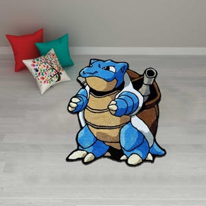 Könnte beinhalten: Blau-beiger Blastoise-Teppich, ein Pokémon-Charakter, mit braunem Panzer und zwei Kanonen. Der Teppich liegt auf einem hellen Holzboden, mit roten, türkisfarbenen und einem kissen mit Baummotiv im Hintergrund.