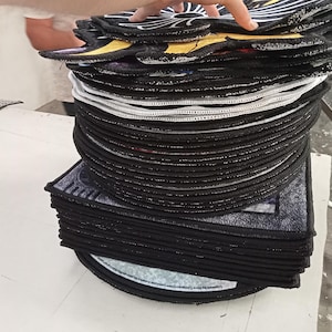 Pode incluir: Uma pilha de tapetes decorativos pretos e redondos com costuras brancas nas bordas. Os tapetes t&ecirc;m um desenho na superf&iacute;cie superior com detalhes em amarelo e branco. Os tapetes est&atilde;o empilhados uns sobre os outros.