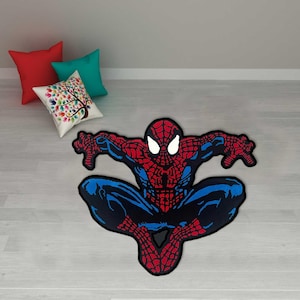 Puede incluir: Una alfombra de Spider-Man roja, azul y negra en pose agachada. La alfombra presenta el diseño icónico del disfraz con detalles de ojos blancos. La alfombra está sobre un piso de madera clara, con cojines rojos, turquesa y estampados en el fondo.