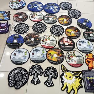 Pode incluir: Uma cole&ccedil;&atilde;o de v&aacute;rios tapetes redondos e com formato, incluindo designs de jogos PlayStation 2, logotipos Chrome Hearts e personagens Pok&eacute;mon. Os tapetes s&atilde;o em v&aacute;rias cores, incluindo azul, preto, branco e laranja.