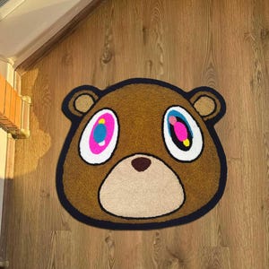 Pode incluir: Tapete em forma de urso castanho com uma borda preta. O urso tem olhos grandes e coloridos com detalhes em rosa, azul e amarelo. O tapete est&aacute; colocado sobre um ch&atilde;o de madeira, adicionando um toque l&uacute;dico.