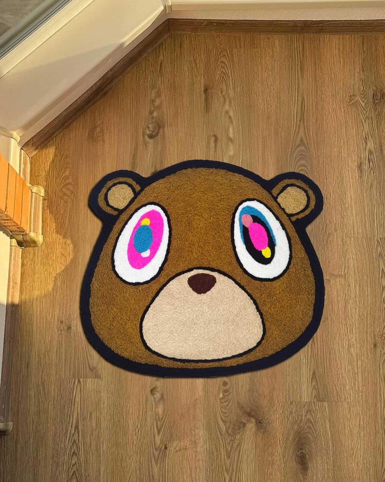 Pode incluir: Tapete em forma de urso castanho com uma borda preta. O urso tem olhos grandes e coloridos com detalhes em rosa, azul e amarelo. O tapete est&aacute; colocado sobre um ch&atilde;o de madeira, adicionando um toque l&uacute;dico.