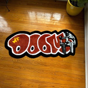 Puede incluir: Una alfombra rectangular con la palabra "DOOM" en una fuente estilizada. Las letras son de color marrón rojizo con contornos blancos y un borde negro. La alfombra también presenta un gráfico de una máscara.