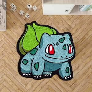 Peut inclure: Tapis en forme de Bulbasaur bleu et vert, avec des yeux rouges et des taches vertes. Le tapis a un contour noir et est posé sur un sol en bois. Un article de décoration.