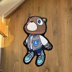 Pode incluir: Um tapete em forma de urso castanho com um rosto branco e uma jaqueta universit&aacute;ria. A jaqueta &eacute; cinza com um "U" azul na frente. O urso est&aacute; usando jeans azuis e t&ecirc;nis azul claro. O tapete est&aacute; em um piso de madeira.