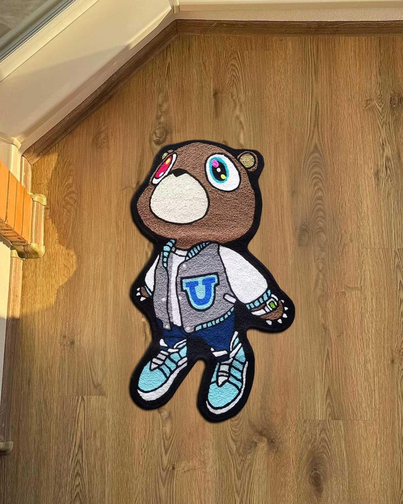 Pode incluir: Um tapete em forma de urso castanho com um rosto branco e uma jaqueta universit&aacute;ria. A jaqueta &eacute; cinza com um "U" azul na frente. O urso est&aacute; usando jeans azuis e t&ecirc;nis azul claro. O tapete est&aacute; em um piso de madeira.