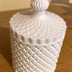 Könnte beinhalten: Ein weißes Keramikgefäß mit Deckel. Das Gefäß hat ein strukturiertes, geometrisches Muster auf Körper und Deckel. Der Deckel ist mit einer dekorativen Spitze versehen. Das Gefäß steht auf einer Holzoberfläche.