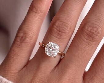 Op de afbeelding: Een solitair ring met een heldere edelsteen in kussenslijpvorm, gezet op een dunne, gepolijste roségouden band. De ring wordt om een vinger gedragen en toont de schittering van de gefacetteerde steen en de gladde, metalen afwerking van de band.