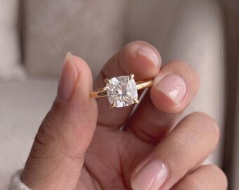 Op de afbeelding: Een gouden ring met een enkele, in kussenvorm geslepen heldere edelsteen die door vier pootjes op zijn plaats wordt gehouden. De ringband is glad en gepolijst en heeft een klassiek ontwerp met een opvallende, gefacetteerde steen die licht reflecteert.