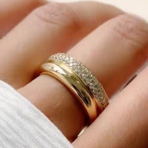 May include: A gold ring featuring a dual-band design. One band is a smooth, polished gold surface, while the adjacent band is encrusted with a dense row of small, clear, sparkling stones that catch the light, creating a layered appearance.