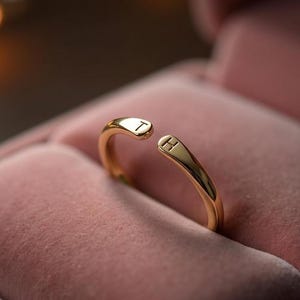 Puede incluir: Un anillo abierto de color dorado con las letras "T" y "H" grabadas en los extremos. El anillo se muestra dentro de un joyero de terciopelo rosa, con una iluminación suave en el fondo. El anillo es una joya delicada.