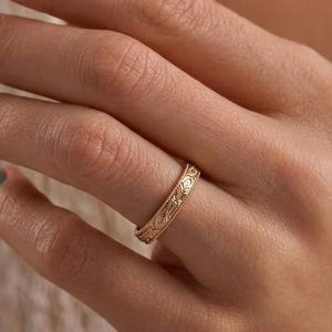 以下が含まれることがあります： 小さな形が複雑に組み合わされた模様のゴールドリング。指にはめられています。リングは金製で、詳細なデザインが施されているようです。背景はぼやけています。リングのデザインが見えます。