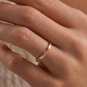 May include: A gold open-style ring featuring a smooth, polished band with a small gap between the two rounded ends. The ring is worn on the middle finger, showcasing a simple, continuous metal design with a reflective, high-shine finish.