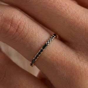 以下が含まれることがあります： ゴールドの細いバンドリングで、バンドの上半分に沿ってバゲットカットの黒い石と小さな丸い黒い石が交互に並んでいます。リングは光沢のある金属仕上げで、指にフィットするスリムなプロファイルが特徴です。