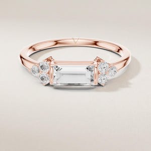 May include: Rose gold ring featuring a rectangular, clear gemstone center stone, flanked by clusters of smaller, round, clear gemstones. The ring has a delicate band and a modern design, suitable for engagement or everyday wear.