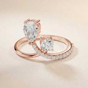 May include: Two rose gold rings with sparkling white gemstones. One ring features a pear-shaped stone, while the other has a round stone and a curved band with smaller stones. The rings are set against a neutral background.