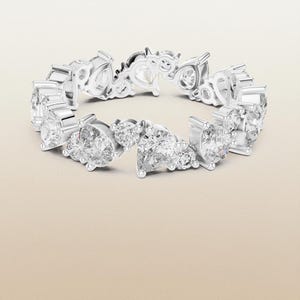 May include: A silver ring adorned with numerous clear, sparkling gemstones. The ring showcases a repeating pattern of pear and round-cut stones, forming a continuous band. The gemstones are set in a silver band, presented against a neutral backdrop.