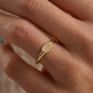Puede incluir: Un anillo de sello dorado con una cara ovalada. El anillo presenta la letra "B" grabada en escritura cursiva. El anillo se muestra en un dedo, centrándose en la joya.
