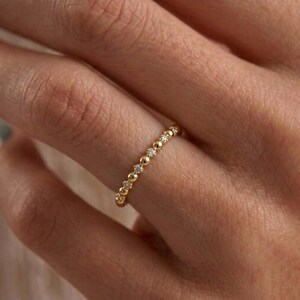May include: A gold ring featuring a delicate band composed of alternating small polished gold spheres and tiny clear round-cut stones set in a continuous row, creating a textured and reflective surface across the finger.
