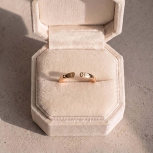 Könnte beinhalten: Ein goldfarbener offener Ring mit zwei flachen, abgerundeten Enden. Ein Ende ist mit dem Buchstaben T und das andere Ende mit dem Buchstaben H graviert. Der Ring befindet sich in einer hellbeigen, mit Samt ausgekleideten, achteckigen Schmuckschatulle.