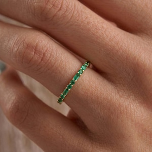 May include: A gold band ring set with emerald-green gemstones. The ring features a continuous row of rectangular-cut stones, creating a vibrant green color. The ring is shown on a person's finger.