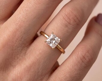 Op de afbeelding: Een gouden ring met een enkele, in princess-geslepen, heldere edelsteen in een zetting met vier pootjes. De ringband is glad en gepolijst en toont een klassiek ontwerp met een vierkante steen die licht reflecteert via het gefacetteerde oppervlak.