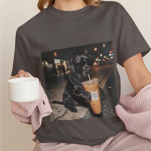 May include: A charcoal grey short-sleeve t-shirt featuring a graphic print of a black Labrador dog wearing sunglasses and holding a plastic cup of iced coffee with a straw. The dog is positioned on a pavement at night with blurred city lights.
