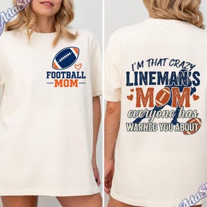Könnte beinhalten: Ein cremefarbenes T-Shirt mit einer kleinen Football-Grafik und dem Text "FOOTBALL MOM" auf der linken Brust. Die Rückseite zeigt eine große Grafik mit dem Text "I'M THAT CRAZY LINEMAN'S MOM everyone has WARNED YOU ABOUT" und Footballs.