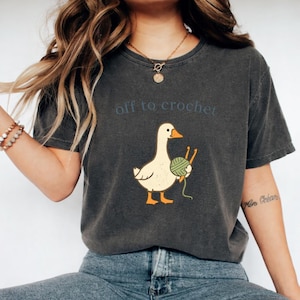 May include: A charcoal gray short-sleeve t-shirt featuring a graphic of a white goose holding a green ball of yarn and two orange crochet hooks. The text "off to crochet" is printed in blue letters above the goose illustration.