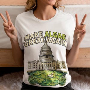 Könnte beinhalten: Ein weißes T-Shirt mit kurzen Ärmeln und einer Grafik des Kapitols der Vereinigten Staaten über einem Teich voller grüner Algen und einer weißen Ente. Der Text "MAKE ALGAE GREAT AGAIN" ist in hellgrünen Buchstaben über dem Gebäude gedruckt.
