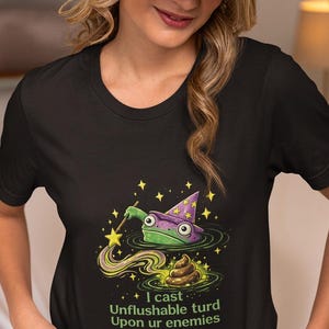 May include: A black t-shirt featuring a graphic of a green frog wearing a purple wizard hat and holding a star-tipped wand. The frog is casting a spell over a brown pile of excrement. The text reads, I cast Unflushable turd Upon ur enemies.