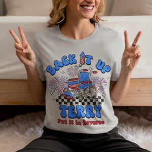 May include: A light gray t-shirt featuring a graphic of a wheelchair with red, white, and blue star and stripe patterns. The text reads BACK IT UP TERRY and Put it in Reverse, accompanied by fireworks, a rocket, and a black and white checkerboard.