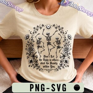 May include: A cream-colored t-shirt featuring a black graphic of three dancing skeletons surrounded by a floral wreath of roses and vines. The text reads, "Don't Let the Ugly in others steal the Beauty within You." A crescent moon sits at the top.
