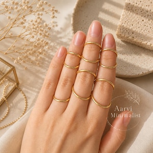 Könnte beinhalten: Ein Set aus acht dünnen, glatten Ringen aus goldfarbenem Metall, die an den Fingern getragen werden. Die Ringe sind schlichte, kreisförmige Bänder mit einer polierten Oberfläche. Der Text Aarvi Minimalist ist unten rechts im Bild zu sehen.