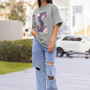 Peut inclure: T-shirt gris avec un motif floral en forme de lettre "S". Le t-shirt est porté avec un jean bleu clair déchiré et des baskets blanches. La personne porte des lunettes de soleil.