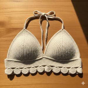 May include: Cream-colored crochet halter top with a scalloped edge. The top features a triangle-shaped bust and a tie-up halter neck. The handmade garment is made of a textured yarn and is displayed on a wooden surface.