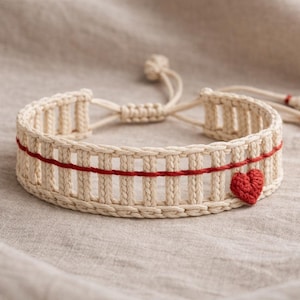 May include: Cream-colored macrame bracelet with a red heart and a red horizontal stripe. The bracelet is adjustable with a sliding knot closure. The macrame design features vertical bars. The bracelet is on a neutral-colored fabric.