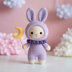 May include: A handmade crochet bunny doll in a lavender outfit with a hood and a ruffled collar. The bunny holds a yellow crescent moon on a wooden stick. The doll has a cream-colored face with rosy cheeks and black eyes.