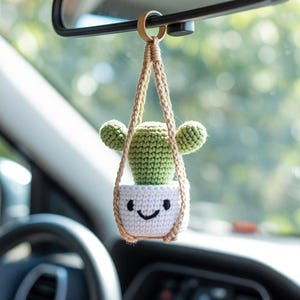May include: A crocheted cactus car charm hanging from a rearview mirror. The charm features a green cactus in a white pot with a smiling face, suspended by a beige macrame hanger. The background is blurred, showing a car interior and a view of the outside.