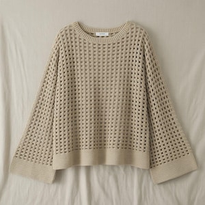 Op de afbeelding: Een beige, open gebreide trui met een ronde hals en lange, uitlopende mouwen. De trui heeft een textuur, rasterachtig patroon en geribde zoom en manchetten. Het kledingstuk wordt op een neutrale achtergrond getoond.